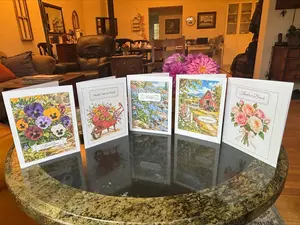 Free printable thank you cards with watercolor pansies, a flower wheelbarrow, forget-me-nots by a stream, a red barn, and a rose bouquet, folded and standing on a table with pink dahlias