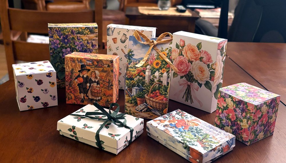 Free printable gift boxes in garden, flower, Thanksgiving, and Christmas designs on a dining table: tall seed packet boxes, square boxes, lid boxes, gift card boxes, and a gable box with a raffia bow