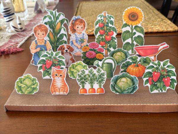 Real paper garden craft standing on a dining room table, with paper corn, a sunflower, tomatoes, peppers, cabbage, lettuce, carrots, a pumpkin, two children, a puppy, and a kitten